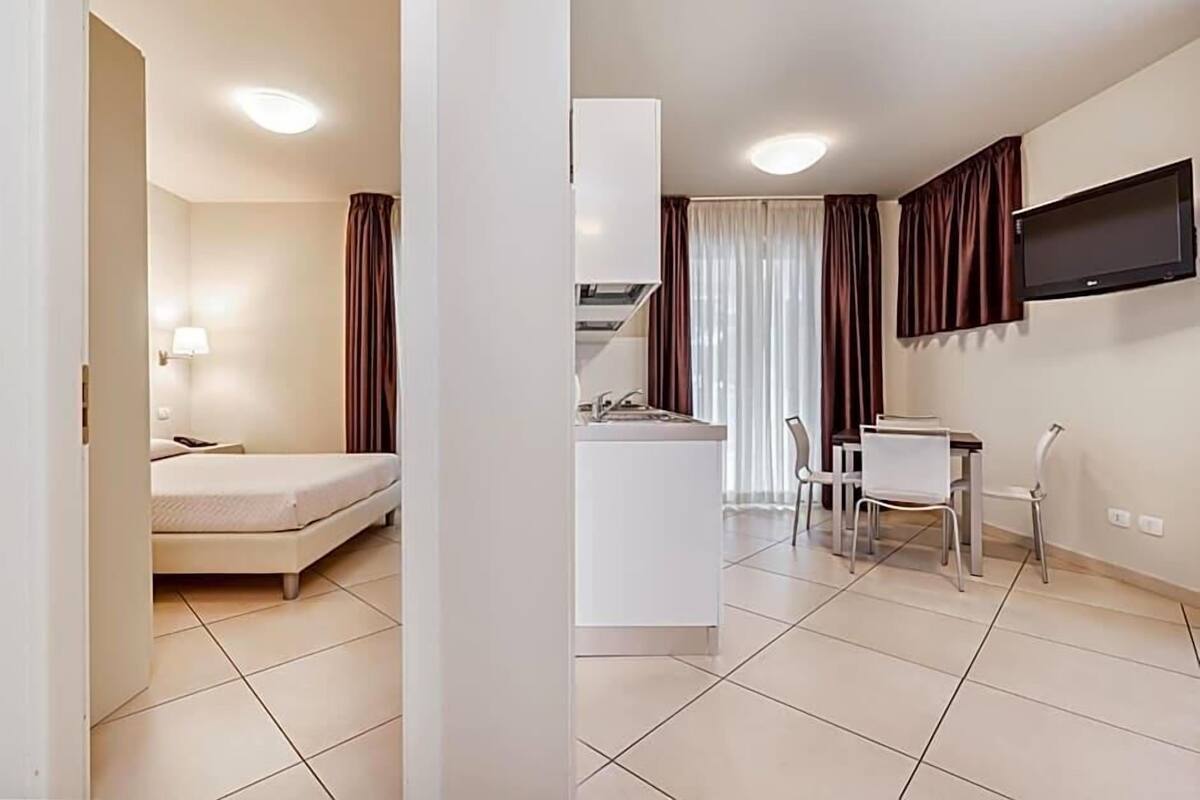 A compact hotel room with a bed, a small kitchenette, a dining area with a table and chairs, and a TV mounted on the wall.