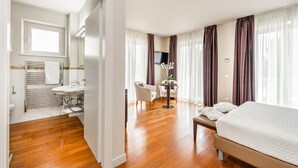 Habitación de hotel con suelo de madera, una cama, un escritorio con silla, televisión y baño con lavamanos y toallero.