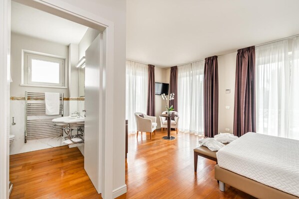 A hotel room with a wooden floor, a bed, a desk with a chair, a TV, and a bathroom with a sink and towel rack.