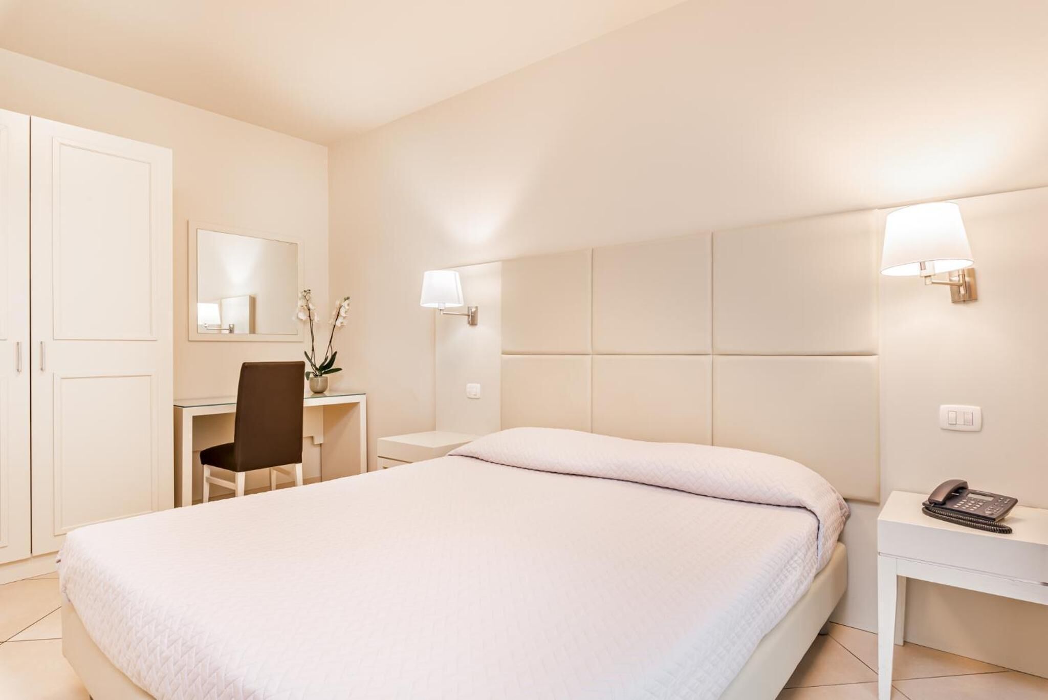 A hotel room with a bed, a desk, a chair, a nightstand with a phone, and a wall-mounted lamp.