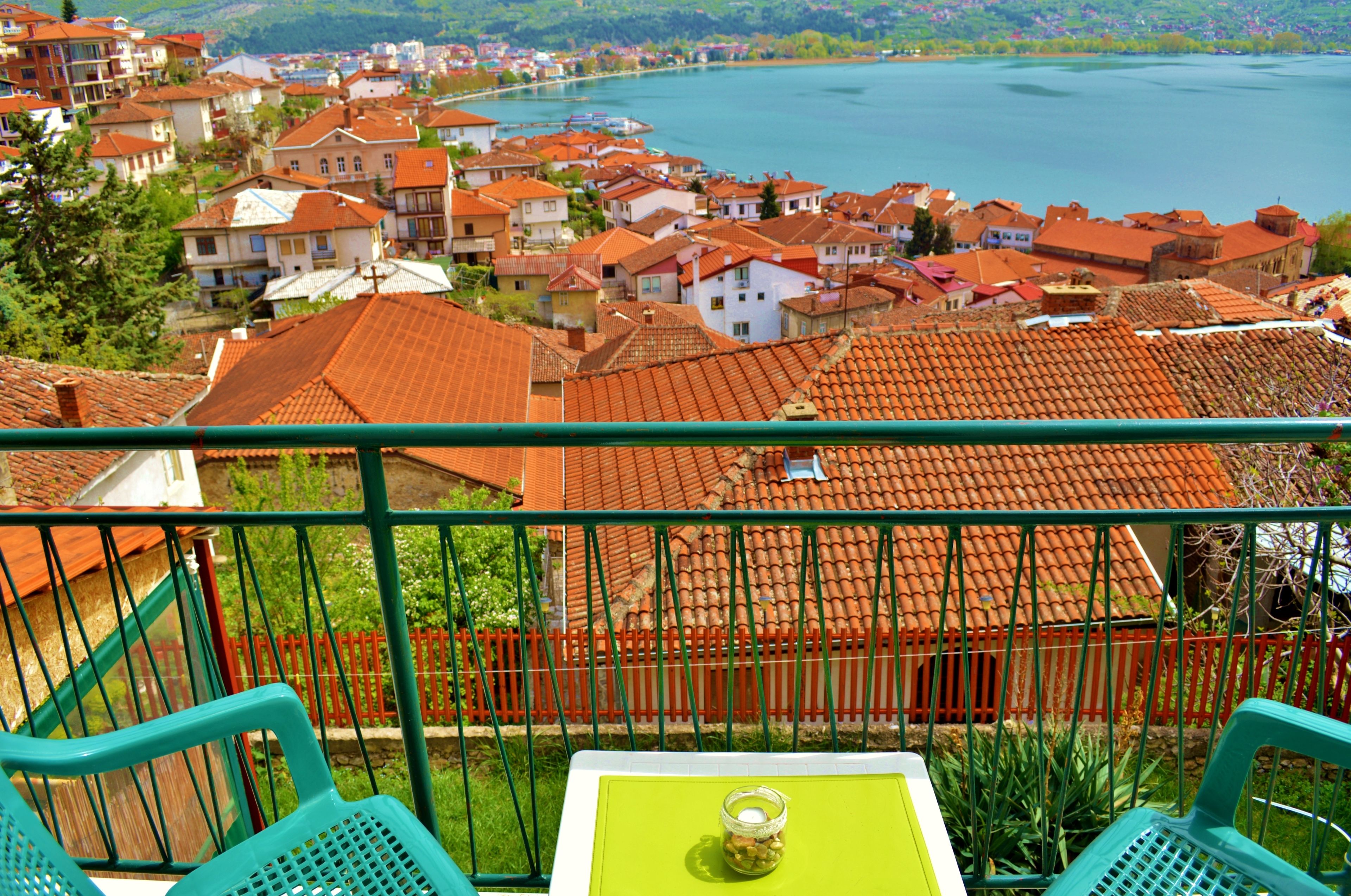 A balcony with green chairs, a table with a glass jar, and a view of a town with red-roofed buildings and a lake.