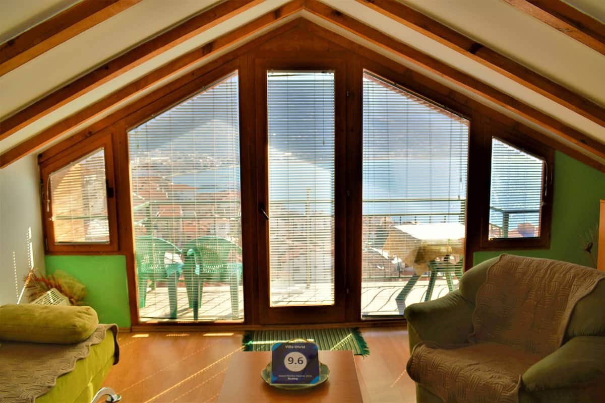 A room with a sloped ceiling, large windows with blinds, a green chair, and a table with a placemat.