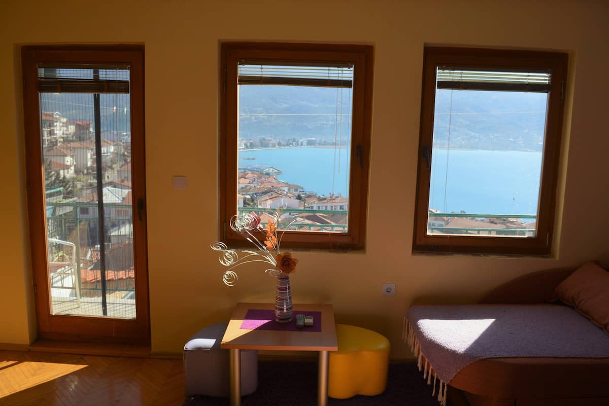 A room with three windows overlooking a lake, a small table with a vase, and a bed with a blanket.