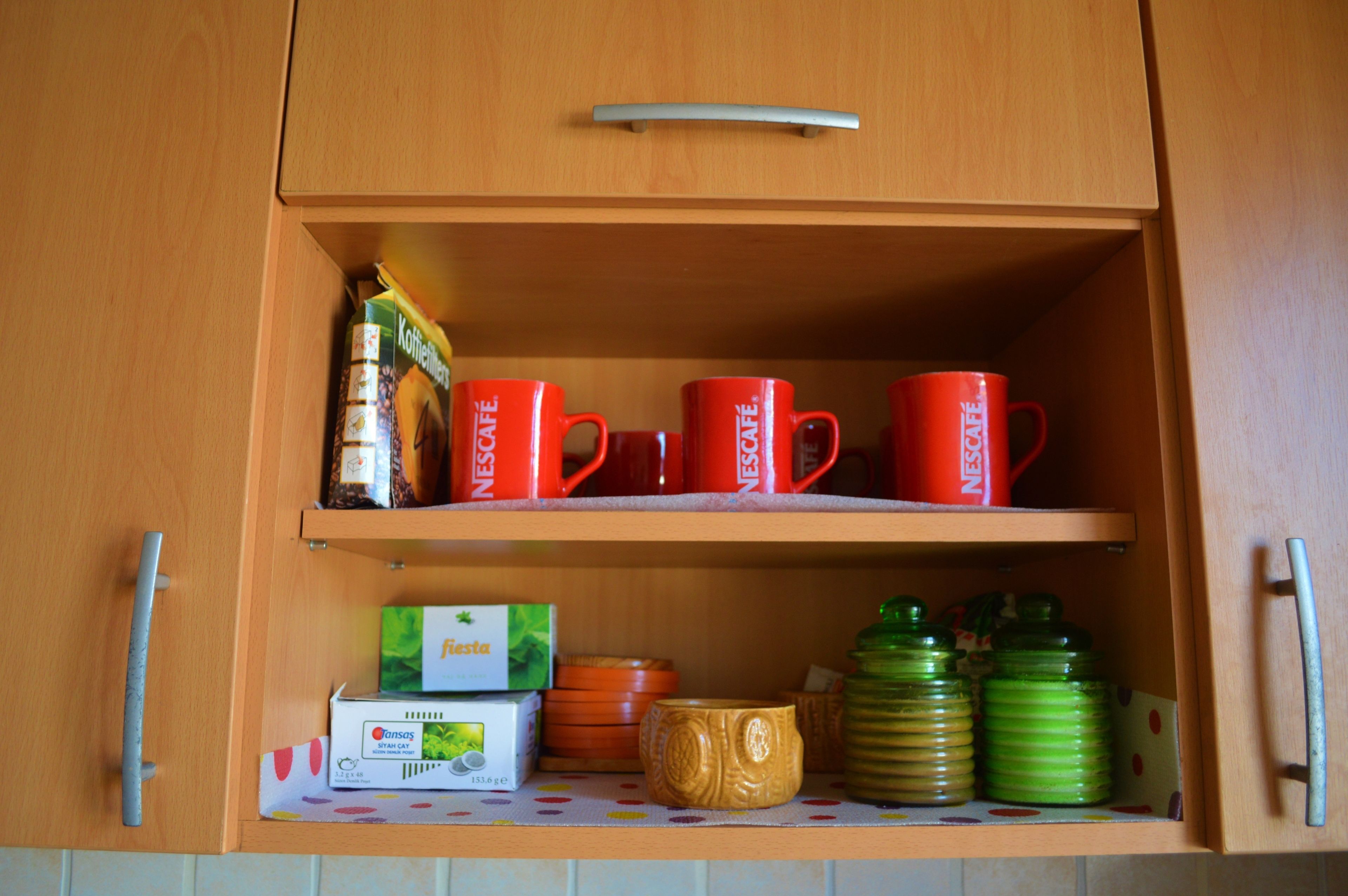 A wooden cabinet with two shelves. The top shelf holds red cups with the word "NESCAFE" and a green box of coffee. The bottom shelf has a green bottle, a yellow mug, and a white box of tea.