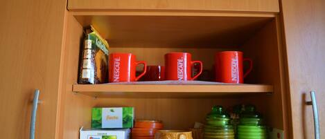A wooden cabinet with two shelves. The top shelf holds red cups with the word "NESCAFE" and a green box of coffee. The bottom shelf has a green bottle, a yellow mug, and a white box of tea.