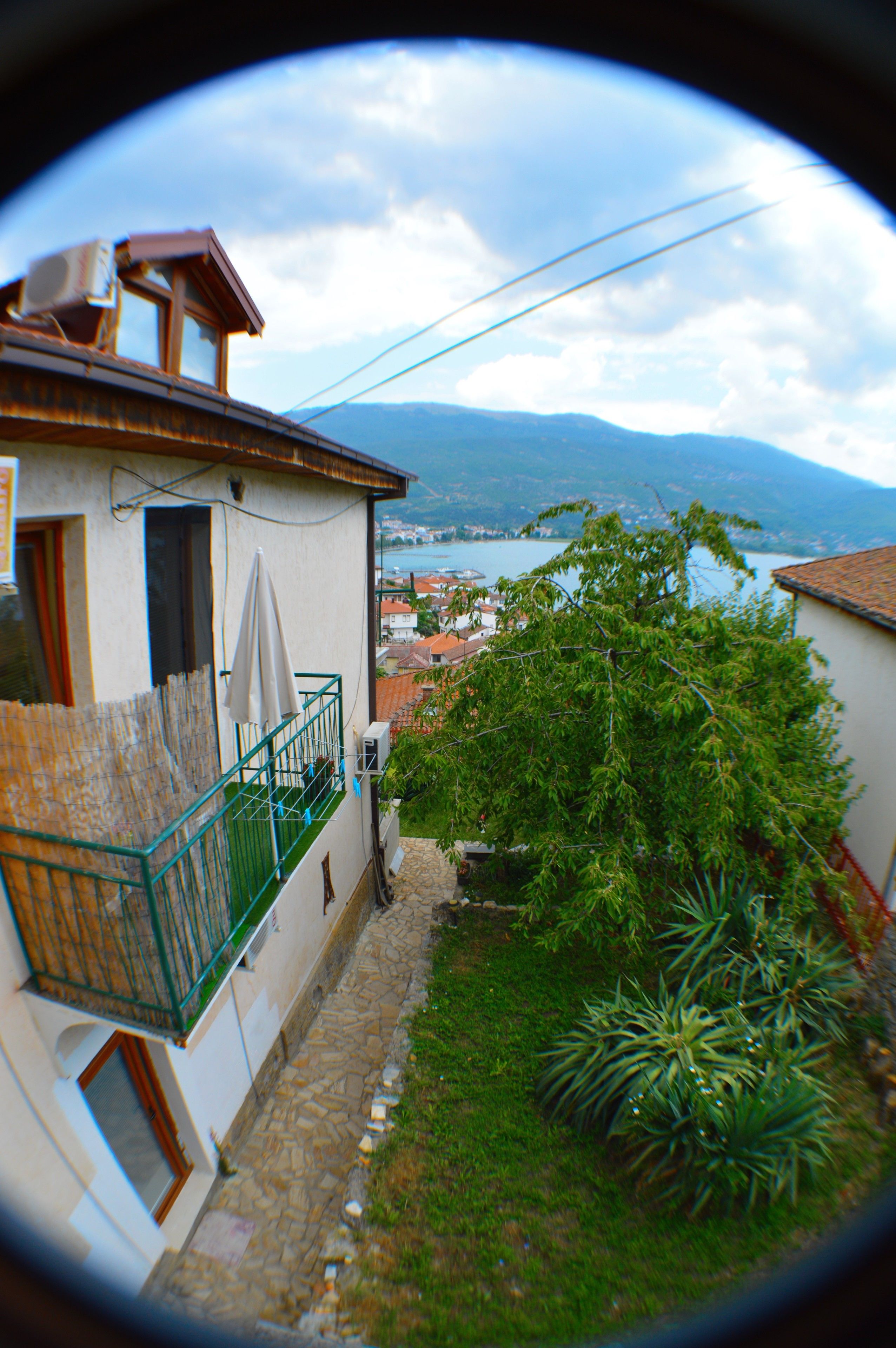 A narrow street with a house on the left, a green railing, and a view of a lake and mountains in the distance.