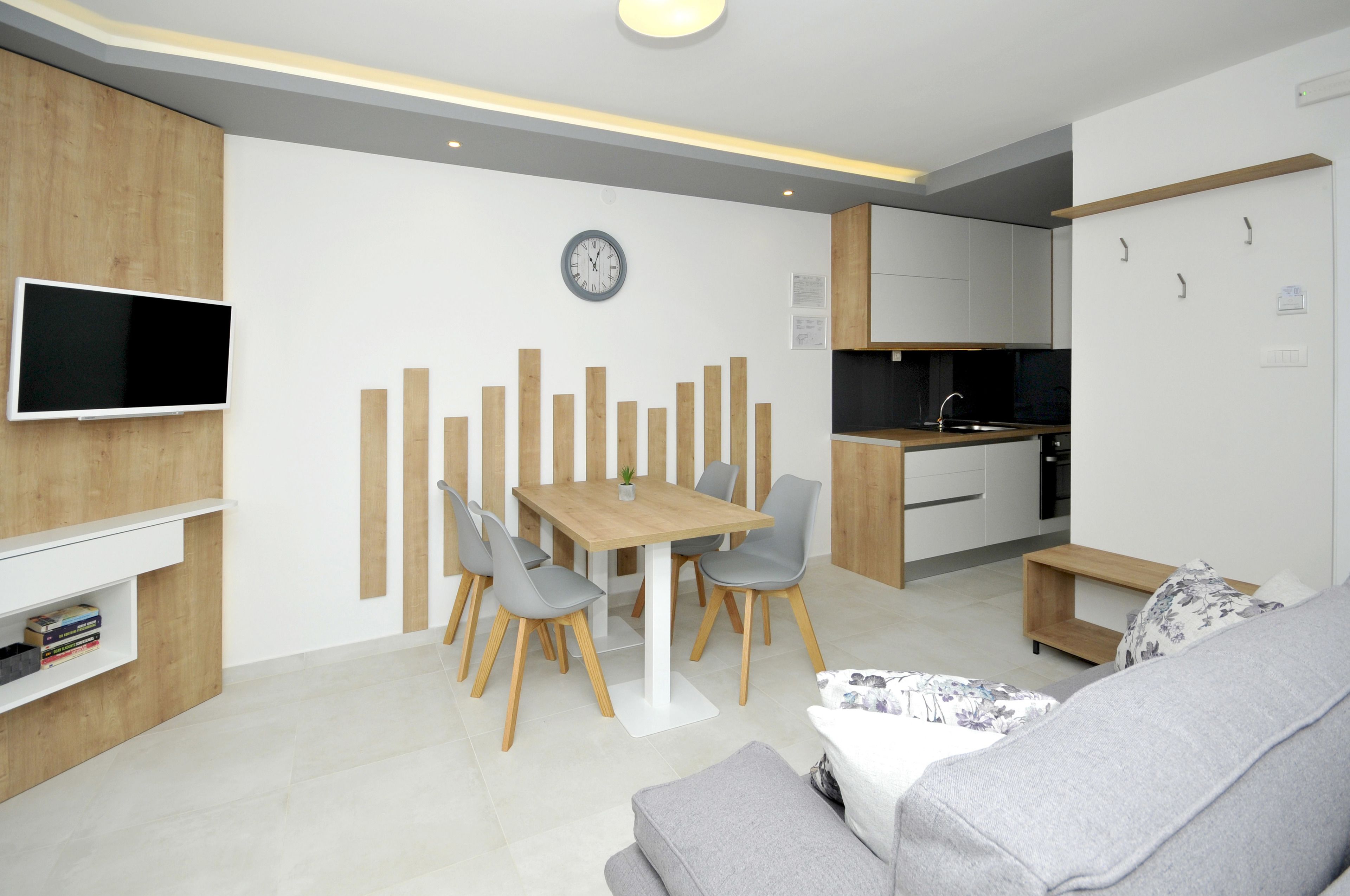 A modern kitchen with a dining area, a flat-screen TV mounted on the wall, and a clock above the table.