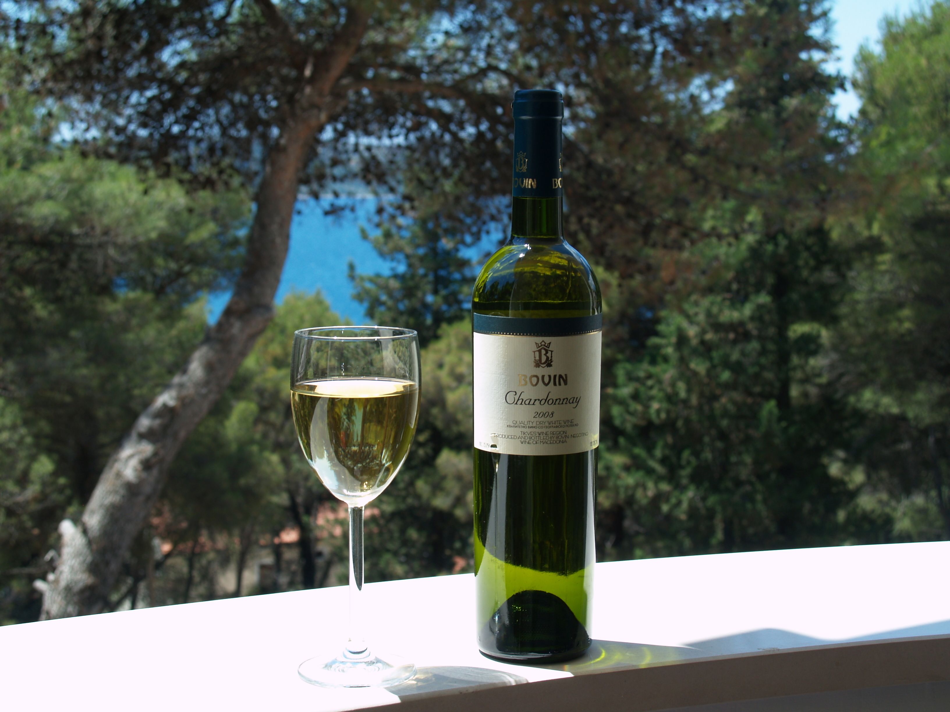 Una bottiglia di vino bianco Chardonnay Bovin 2008 e un bicchiere di vino su una superficie bianca, con uno sfondo naturale all'aperto.