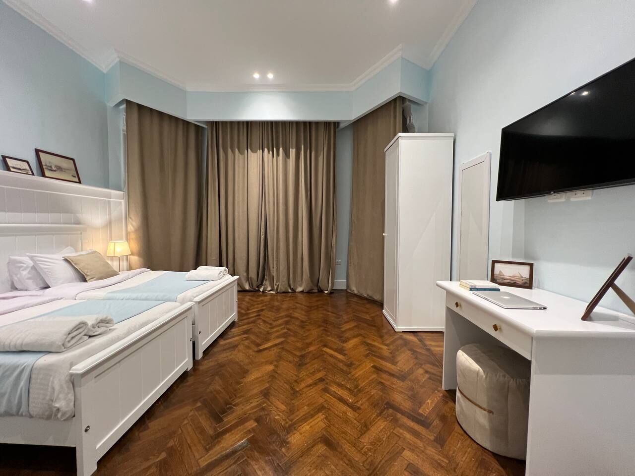 A hotel room with a wooden herringbone floor, a bed with white bedding, a desk with a laptop, and a television mounted on the wall.