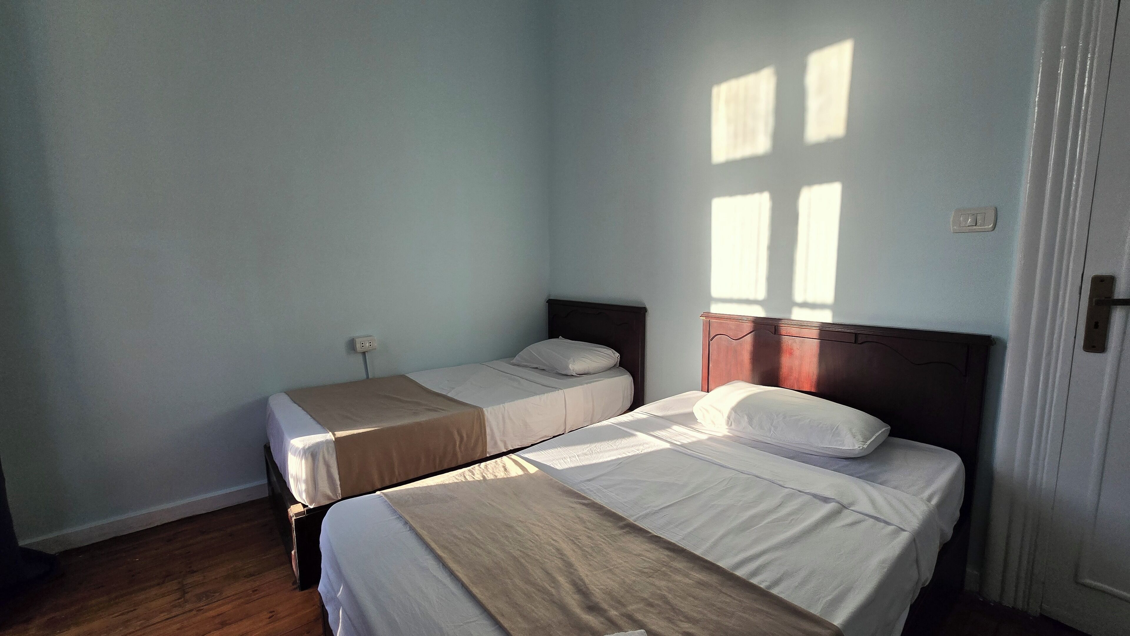 Una habitación con dos camas individuales, suelo de madera, paredes azul claro y una ventana que proyecta sombras.