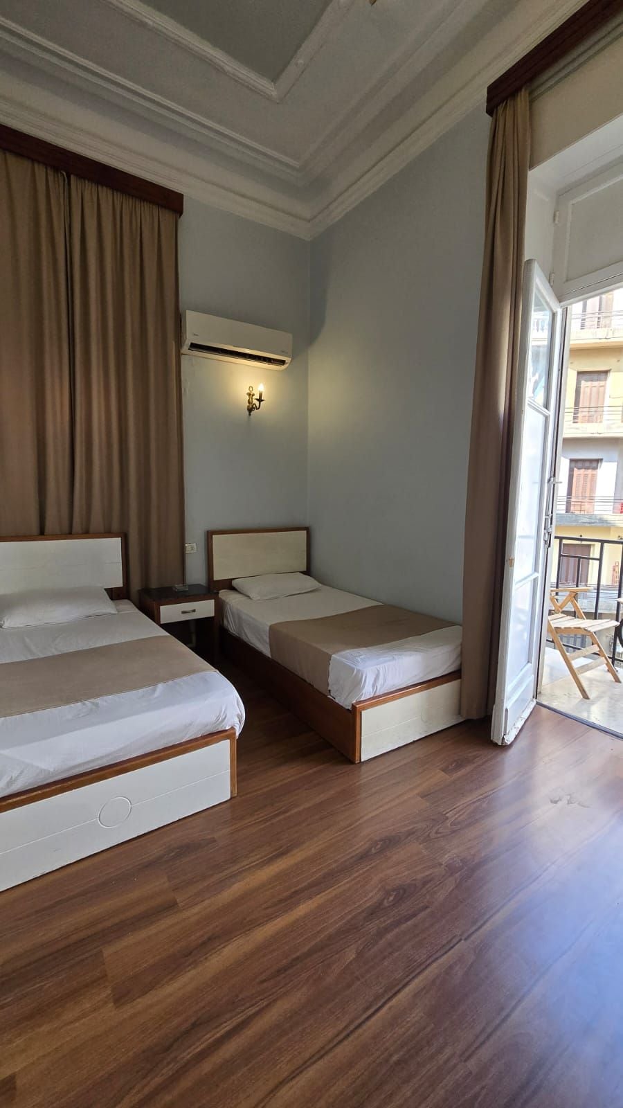 A hotel room with two beds, a nightstand, an air conditioning unit, and a view of the outside balcony.