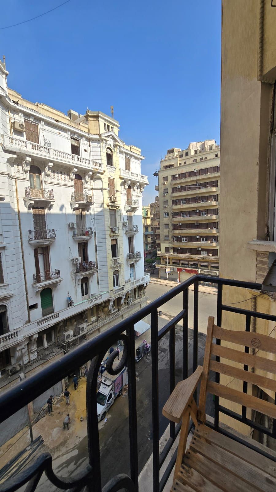 A balcony with a wooden bench, overlooking a street with buildings and a parked vehicle.