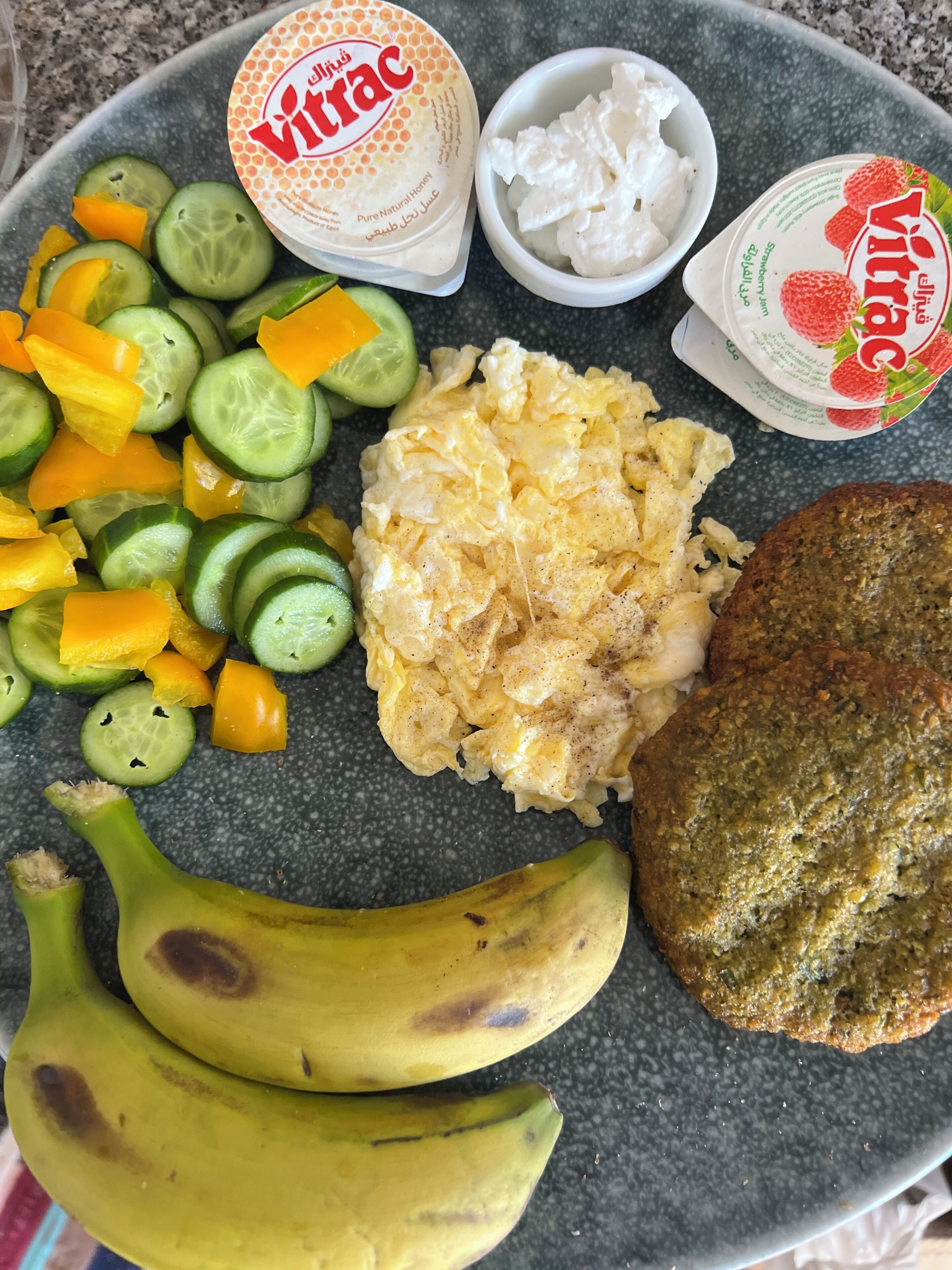 A plate with scrambled eggs, sliced cucumbers, bell peppers, a slice of banana, a piece of toast, and two containers of yogurt.
