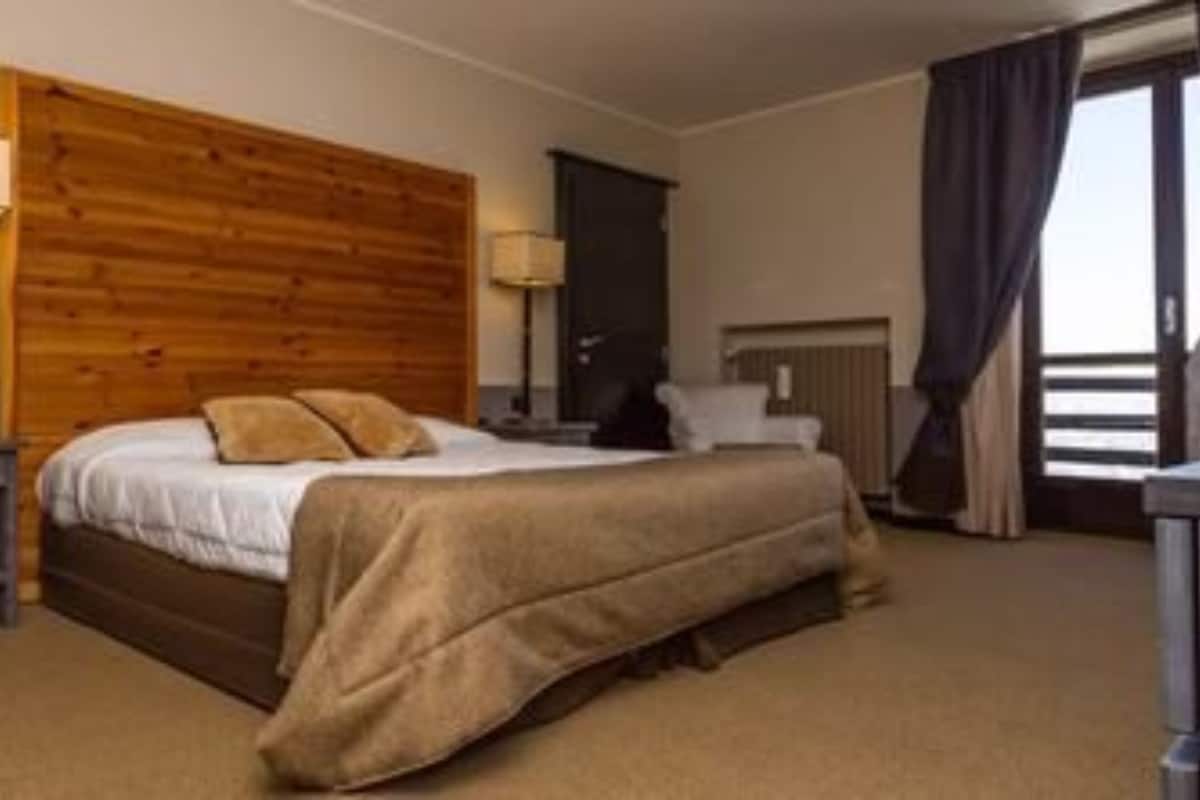 A hotel room with a wooden headboard, a bed with a beige comforter, a bedside table with a lamp, and a view of the outside through a window with curtains.