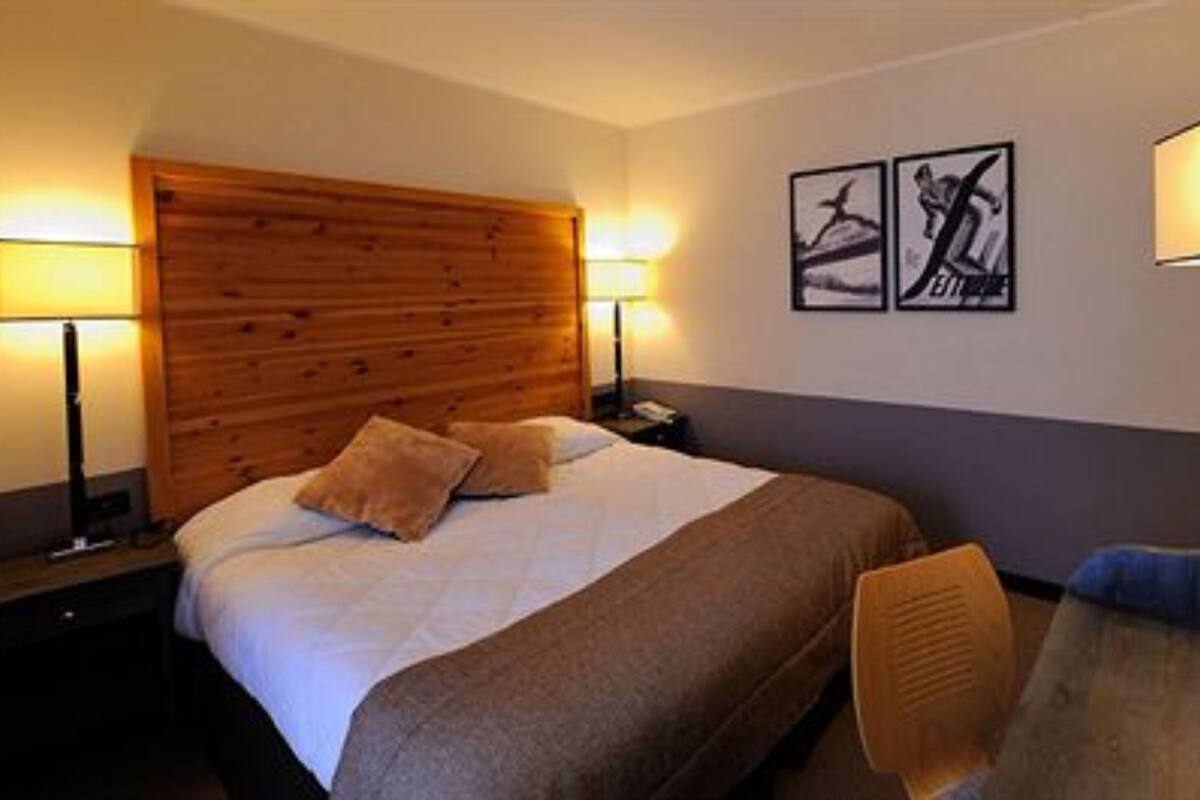 A hotel room with a wooden headboard, a bed with white and brown bedding, two bedside lamps, a desk with a chair, and two framed pictures on the wall.