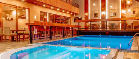 Vestíbulo de hotel con una amplia piscina interior, zona de bar y área de descanso con mesas y sillas.