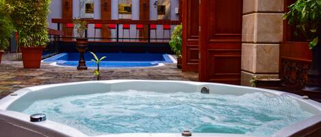 A hot tub in a courtyard with potted plants and a pool in the background.