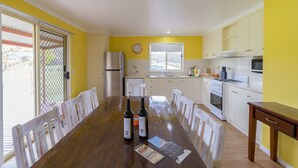 A dining area with a wooden table, chairs, and two bottles of wine. There is a kitchen with white cabinets and a window. - Tokemata Retreat (Cobram East)