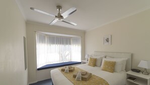 A hotel room with a bed, a window with sheer curtains, a ceiling fan, a bedside table with a lamp, and a small bedside table with a book. - Tokemata Retreat (Cobram East)