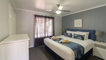 Una habitación de hotel con cama, ventilador de techo, ventana con cortinas, mesita de noche con lámpara y un cuadro colgado en la pared.