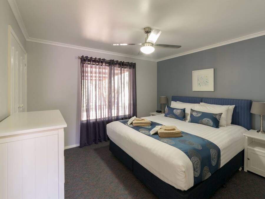 Una habitación de hotel con cama, ventilador de techo, ventana con cortinas, mesita de noche con lámpara y un cuadro colgado en la pared.