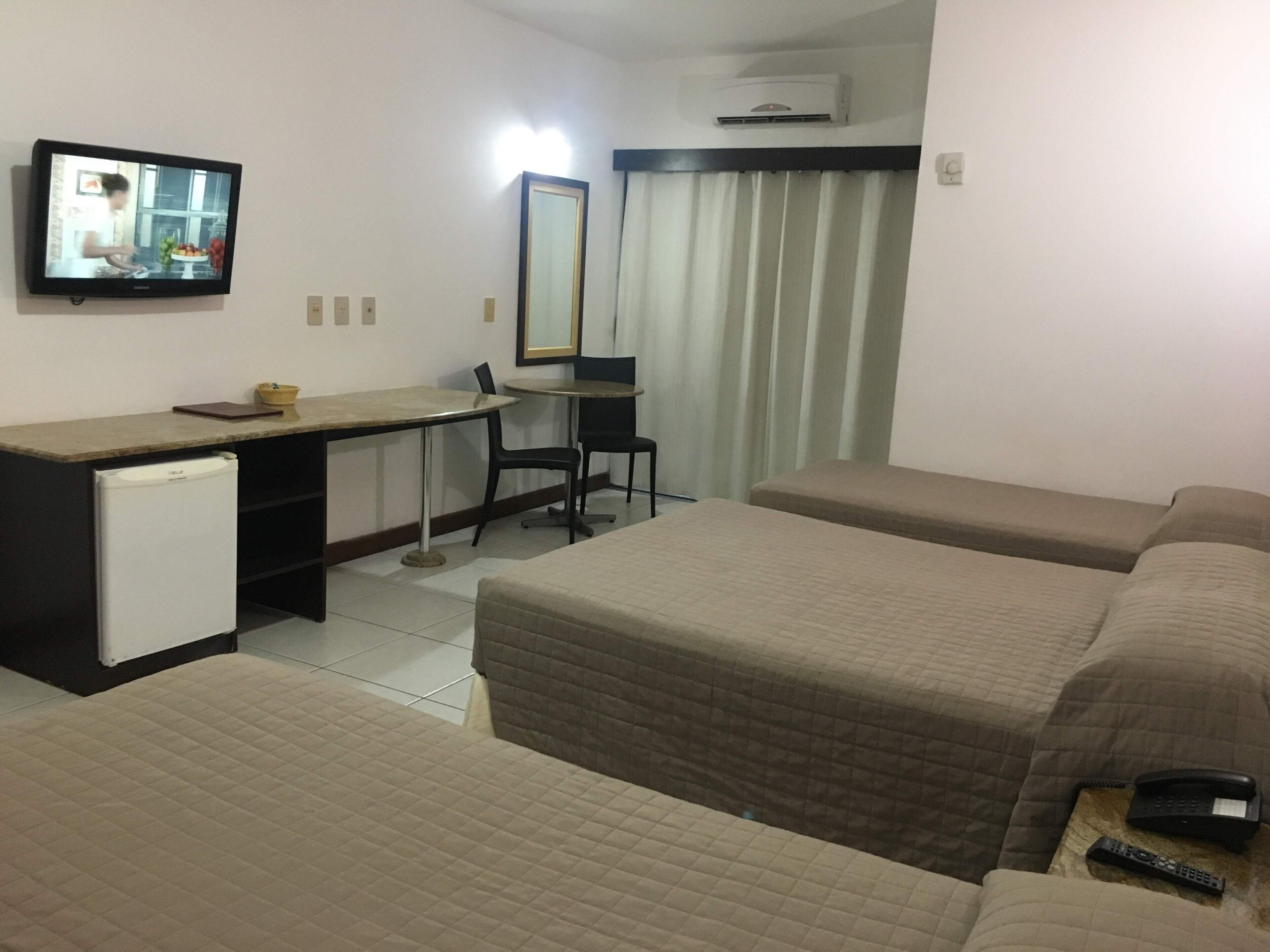 A hotel room with two beds, a desk with a computer, a small fridge, a chair, and a TV displaying a scene.