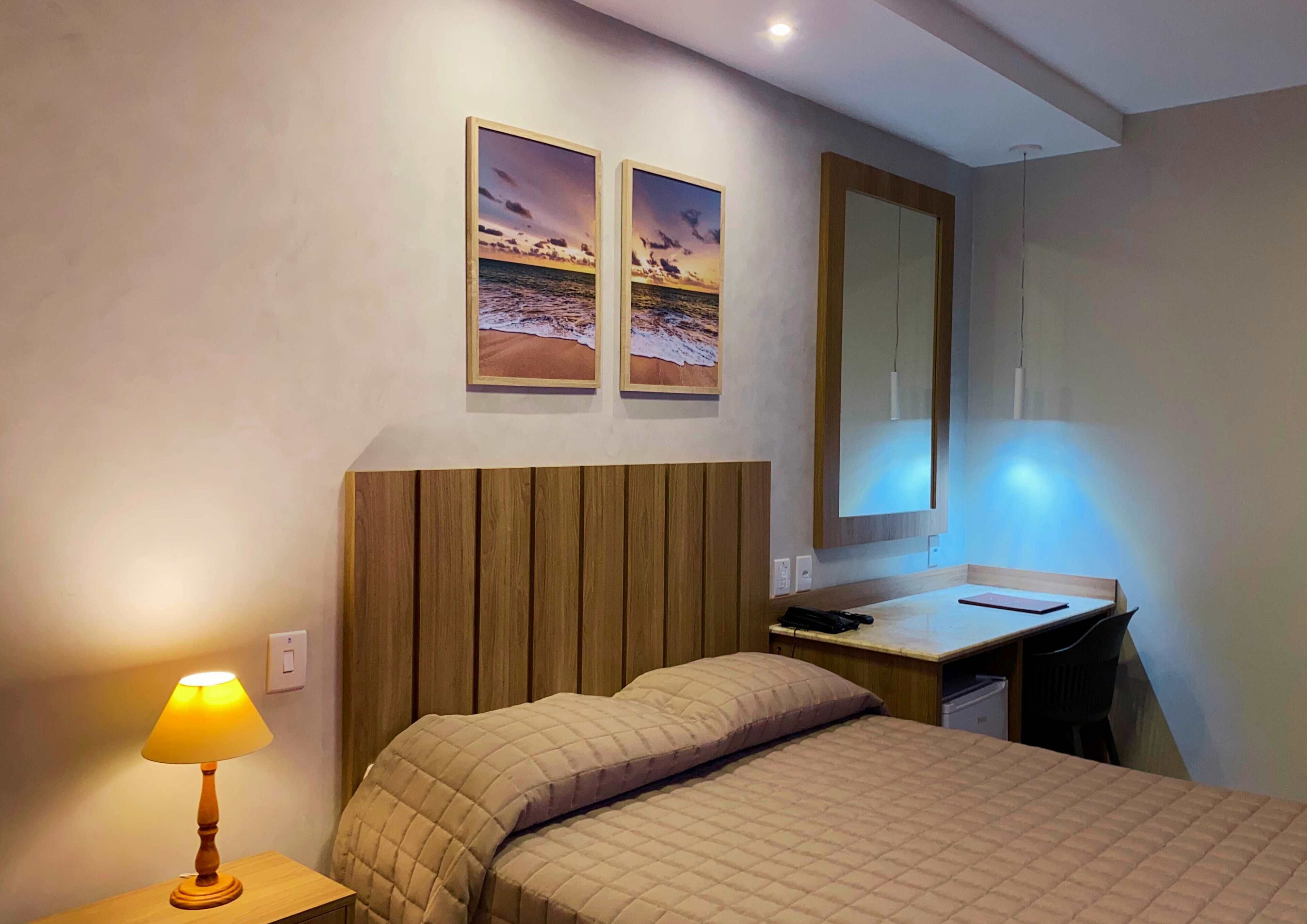 Une chambre d’hôtel avec un lit, un bureau, une lampe et deux tableaux encadrés au mur.