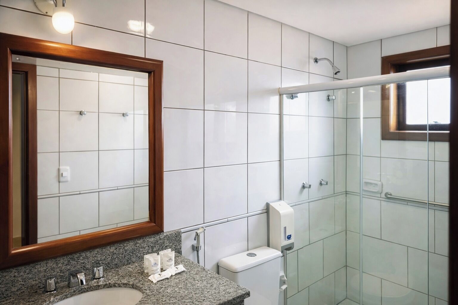 A modern bathroom with a granite countertop, a large mirror, a glass shower enclosure, and a toilet with a paper towel dispenser.