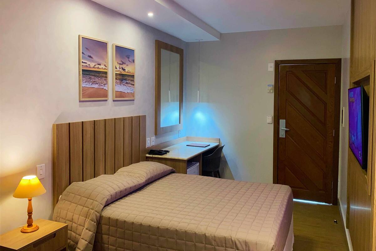 A hotel room with a bed, a desk, a chair, a lamp, a TV, and a door.