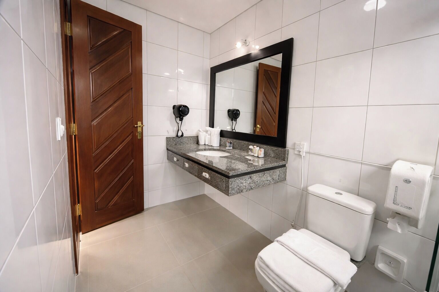 Un baño moderno con puerta de madera, encimera de mármol, espejo grande, inodoro y secador de manos.