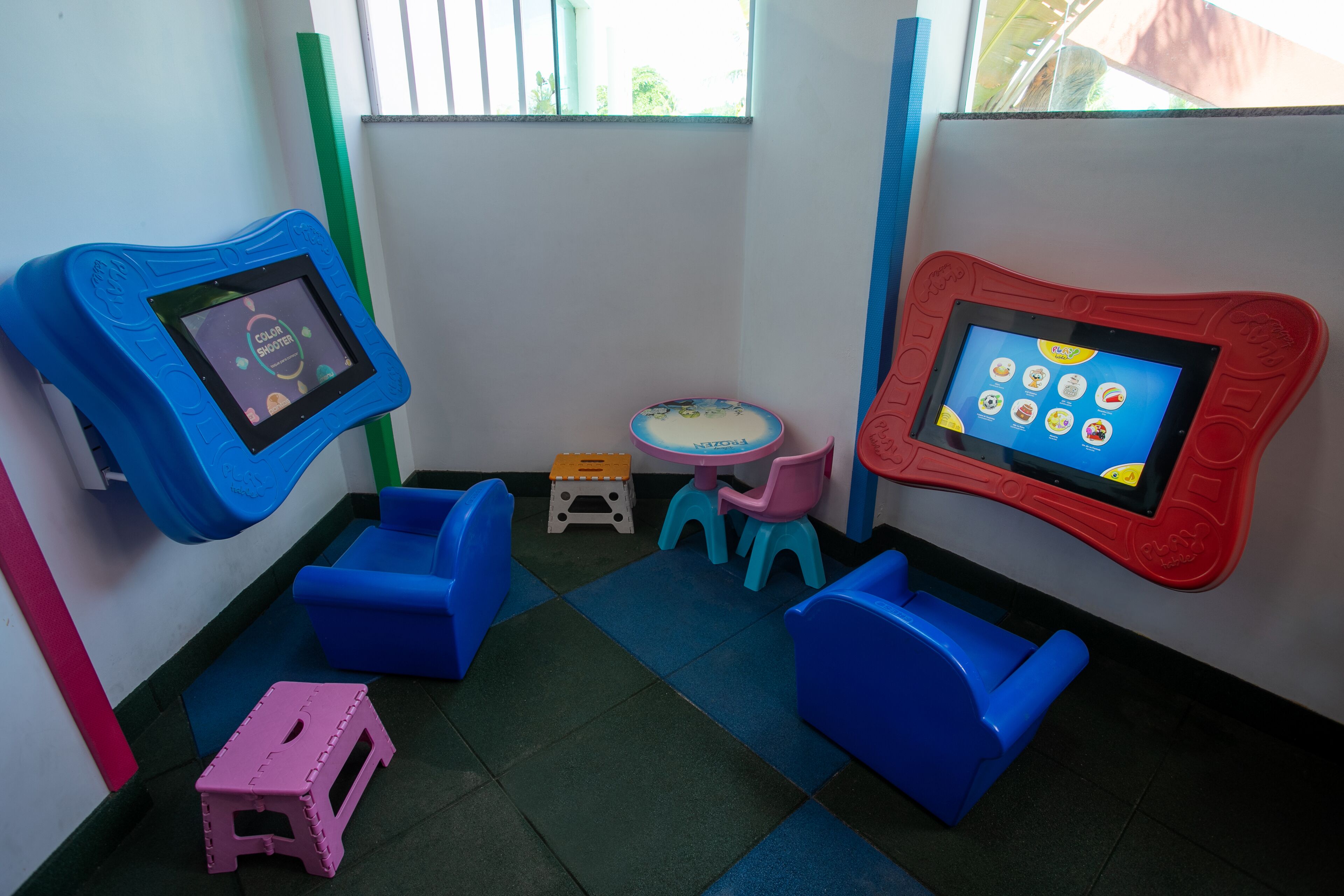 Zona de juegos infantiles con dos estaciones de tabletas, sillas azules y rojas, una mesita y un taburete rosa.