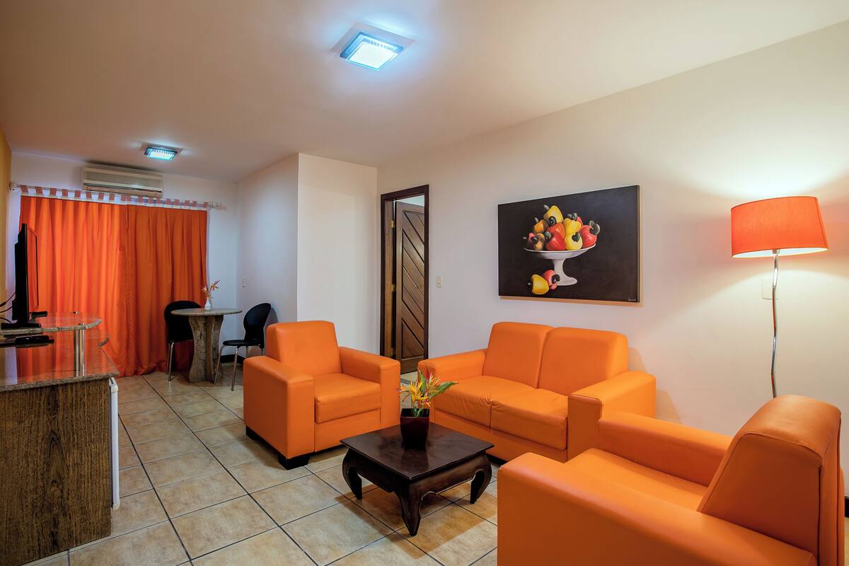 A living room with orange furniture, a coffee table with a plant, a fruit bowl on the wall, and a lamp.