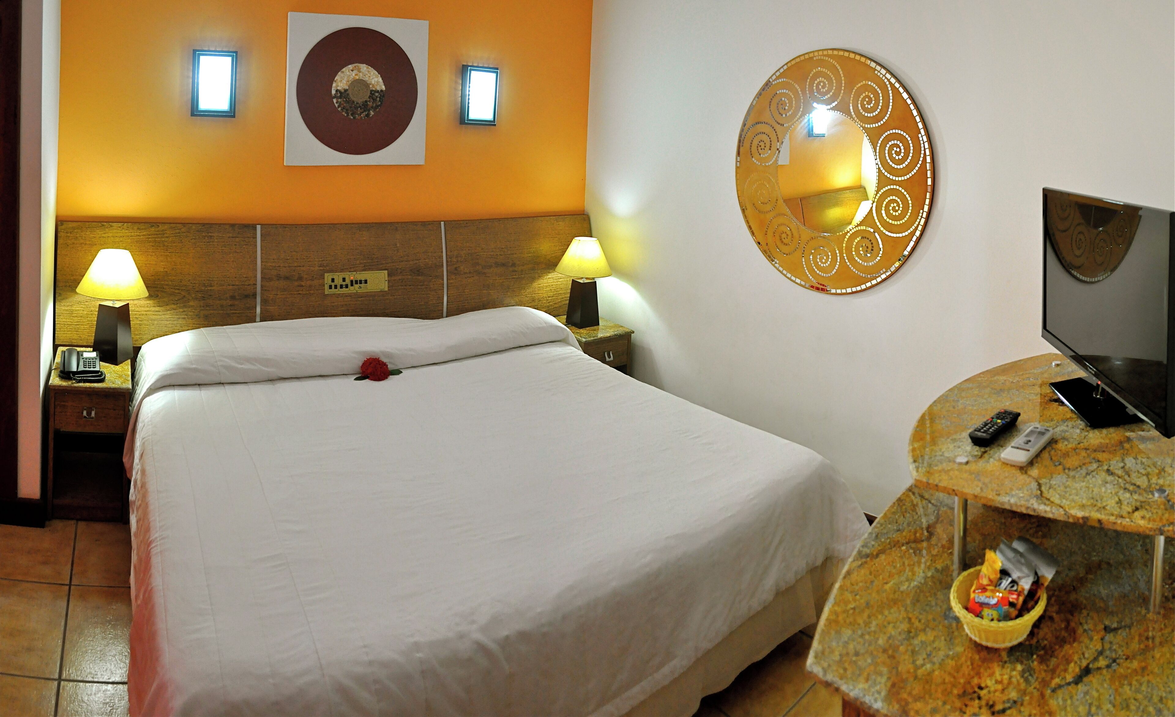 Une chambre d’hôtel comprenant un lit, deux lampes de chevet, un miroir fixé au mur, une télévision et une petite table avec un panier.