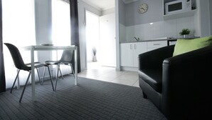 En moderne stue med en sort sofa, et glasbord med to stole, samt et køkkenområde med hvide skabe og en mikrobølgeovn.