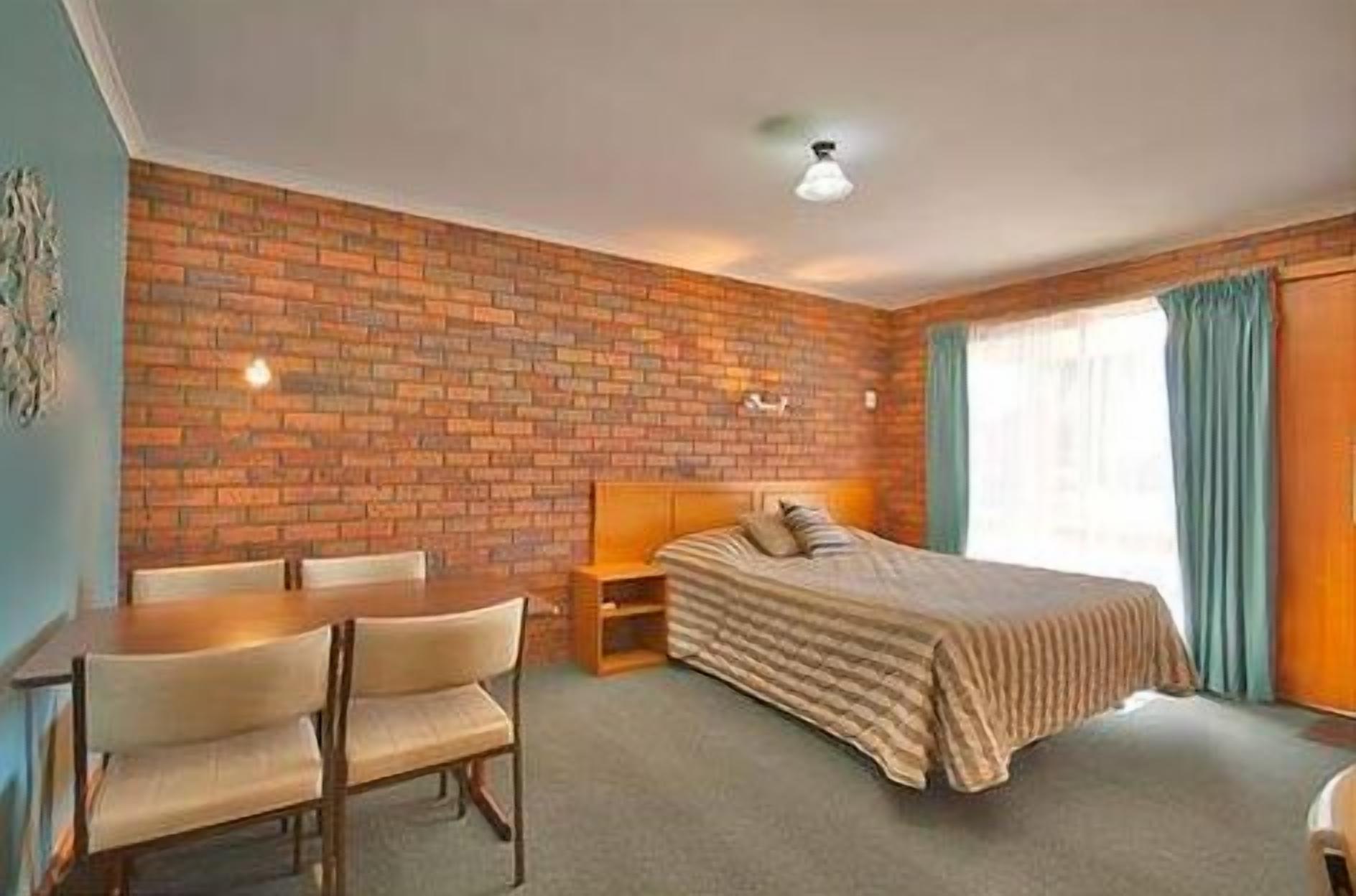 A room with a brick wall, a bed with a striped blanket, a wooden nightstand, a chair, and a table.
