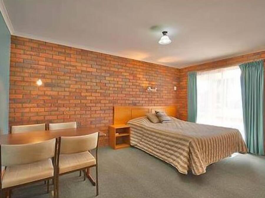 A room with a brick wall, a bed with a striped blanket, a wooden nightstand, a chair, and a table.