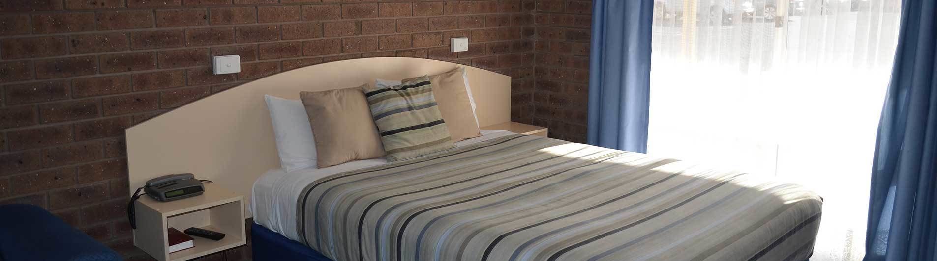 Una camera da letto con parete in mattoni, un letto con biancheria a righe, un comodino con un telefono e una finestra con tende.