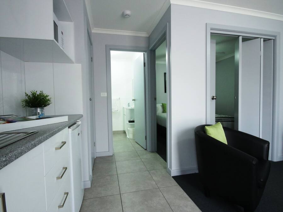 Une cuisine moderne avec des meubles blancs, un comptoir en granit et une petite plante. Un couloir mène à une salle de bain et on y trouve une chaise noire avec un coussin vert.