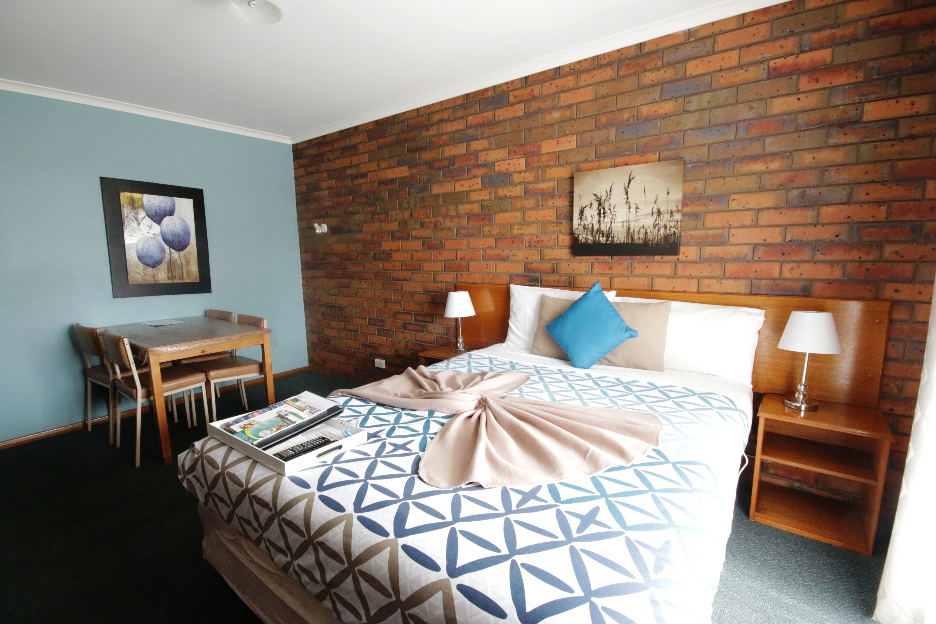 A hotel room with a brick wall, a bed with a geometric patterned bedspread, a desk with a chair, and two bedside lamps.