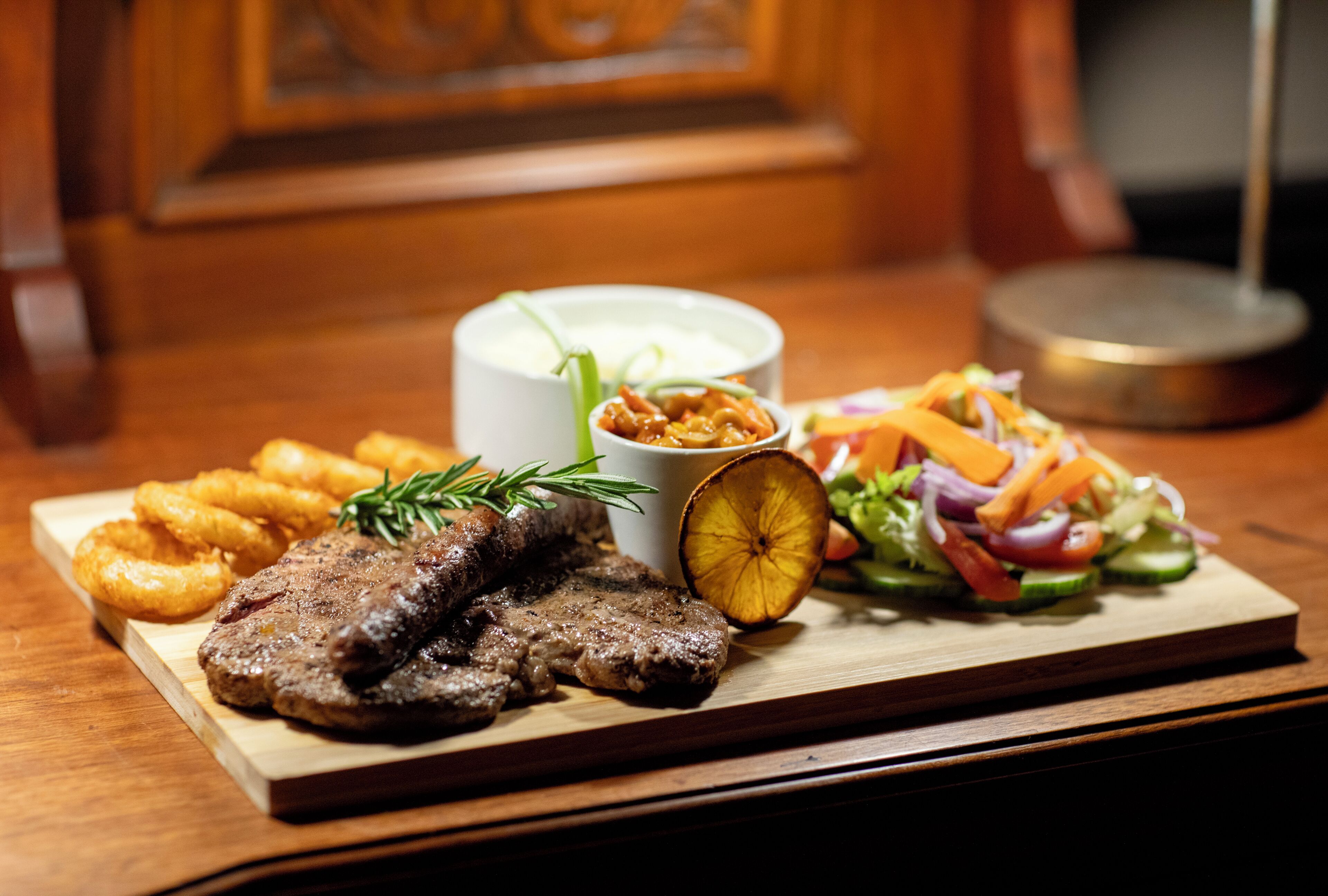 A wooden table with a meal consisting of steak, onion rings, a salad, and a small bowl of sauce.