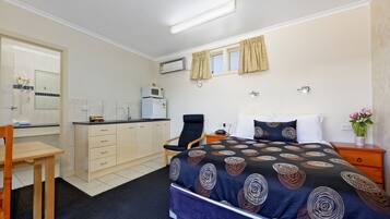 A bedroom with a bed, a desk, a chair, a nightstand, and a small kitchenette.