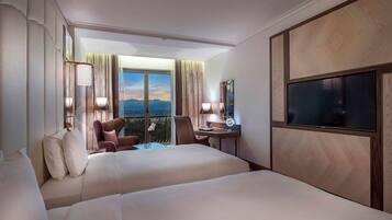 A hotel room with two beds, a desk with a chair, a TV, and a view of the mountains.