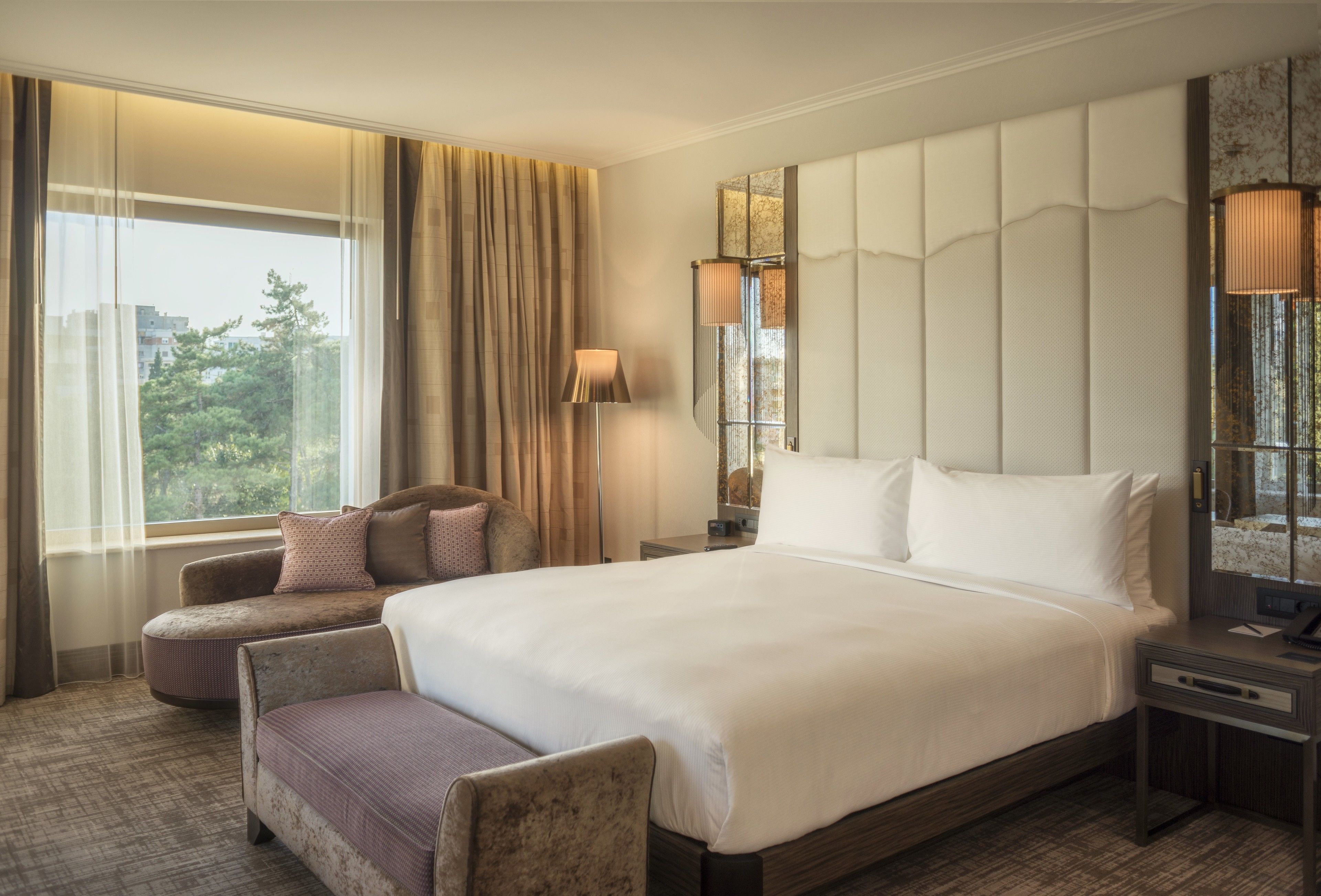 A hotel room with a large bed, two armchairs, a nightstand, a lamp, and a view of trees through the window.