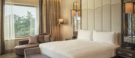 A hotel room with a large bed, two armchairs, a nightstand, a lamp, and a view of trees through the window.