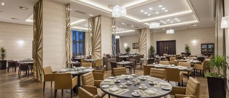 Amplia zona de comedor de hotel con varias mesas redondas dispuestas para comer, rodeada de sillas cómodas.
