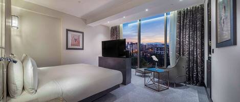 Una camera d'albergo moderna con un letto grande, una TV, un tavolino e una sedia. È presente un'ampia finestra con vista sulla città.