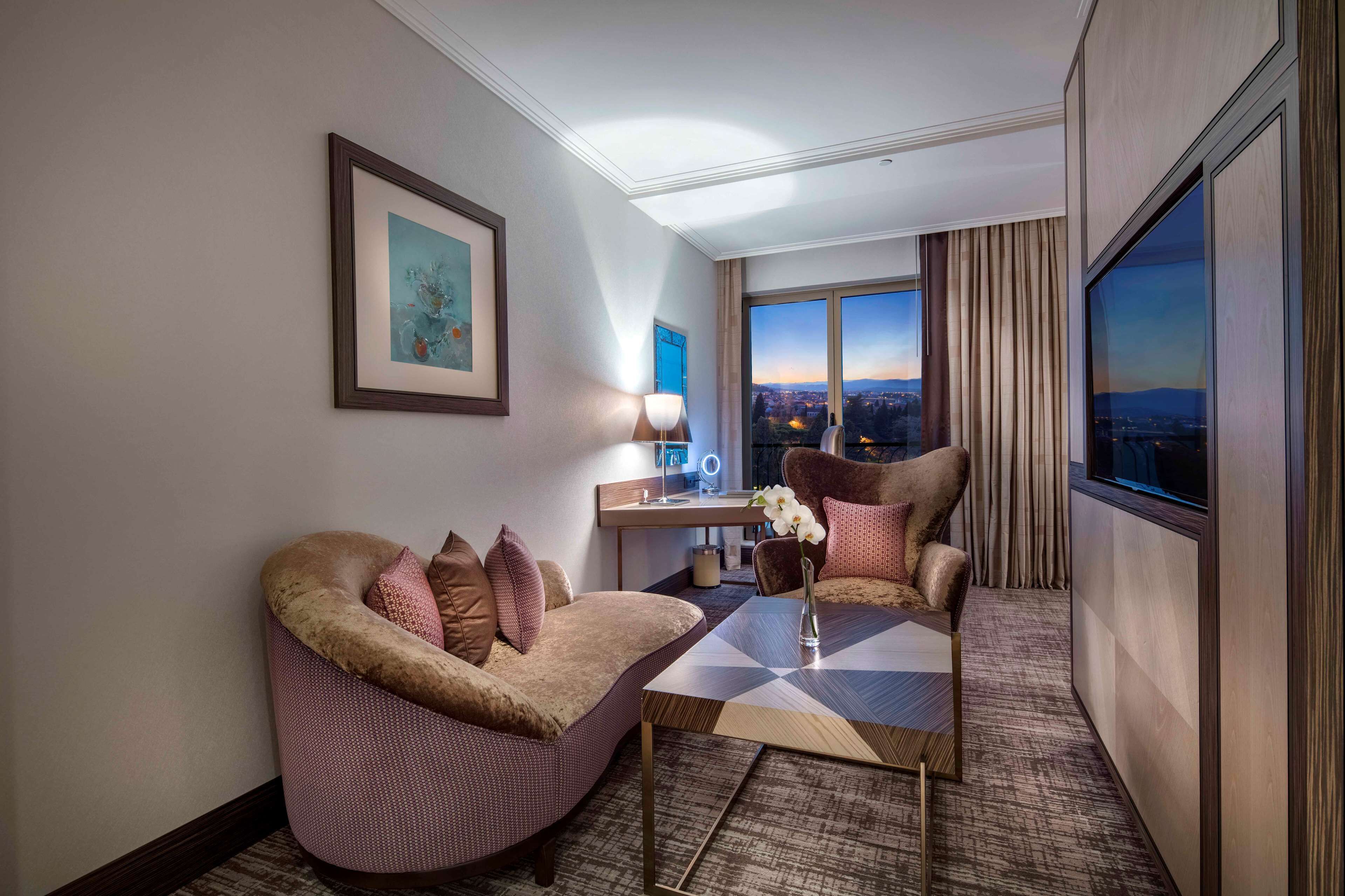A modern hotel room with a curved sofa, a small table, and a view of the cityscape through large windows.