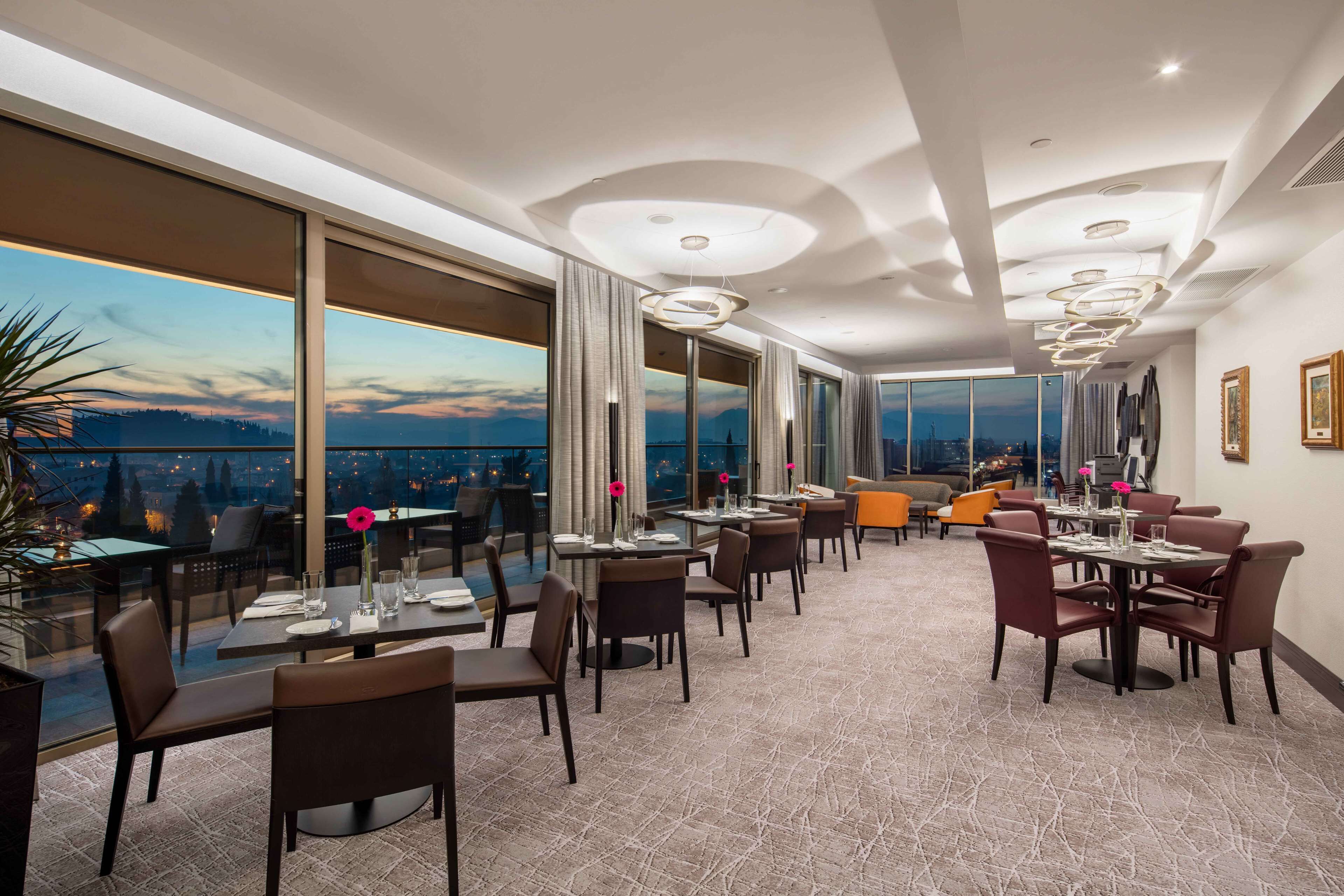 A modern dining area with large windows, a high ceiling with circular light fixtures, and a view of the cityscape at dusk.