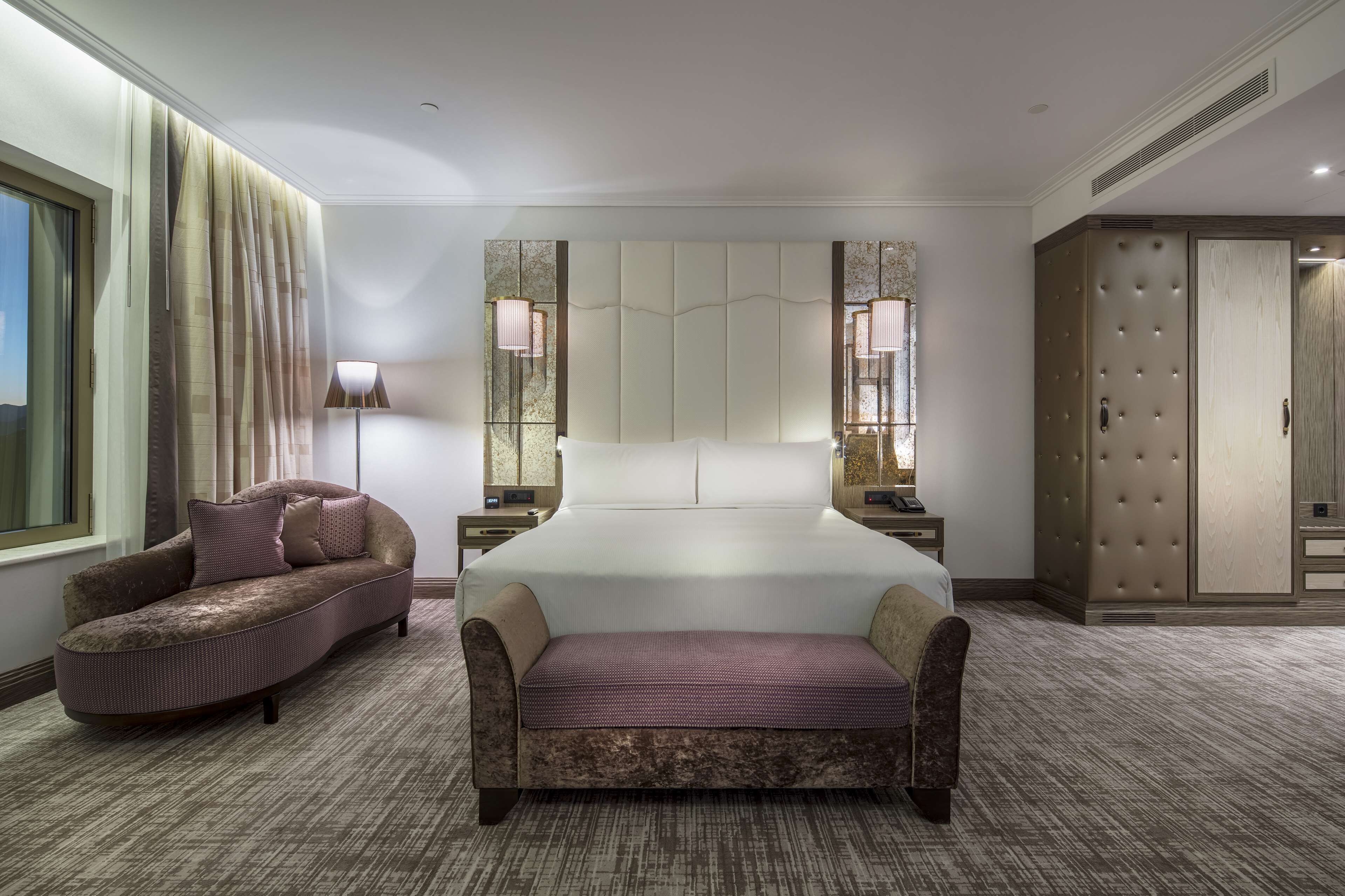 A hotel room with a large bed, a chaise lounge, a wardrobe, and a nightstand with a lamp.