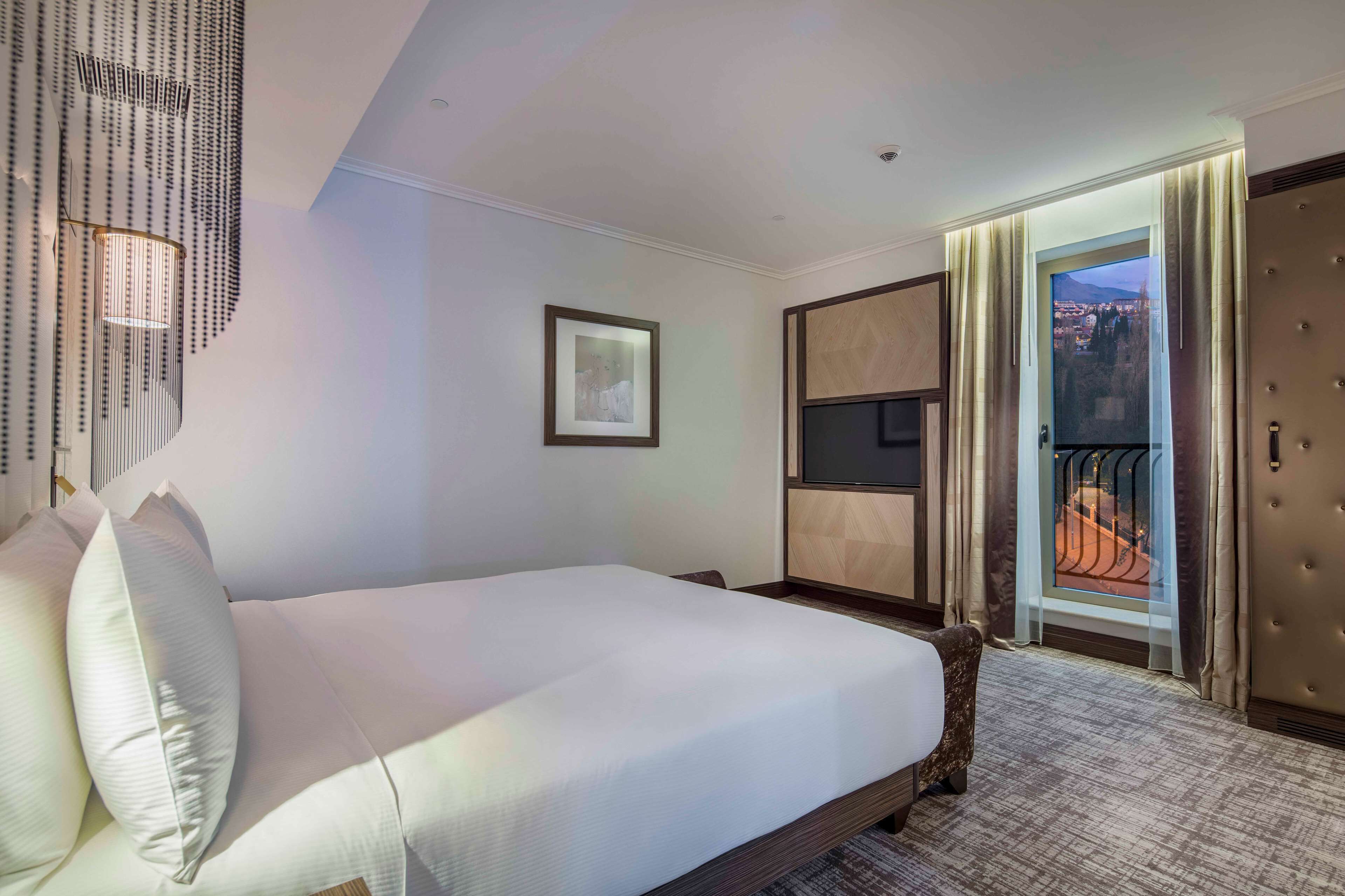 Hotelkamer met een groot bed, een ingelijst schilderij aan de muur en uitzicht op een stadsgezicht door een open deur.